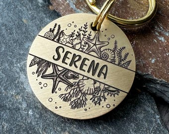 May include: A round, brushed brass metal pet tag featuring a horizontal center band engraved with the name SERENA. The tag is decorated with detailed black line art illustrations of various seashells, starfish, and seaweed, and includes a gold ring.