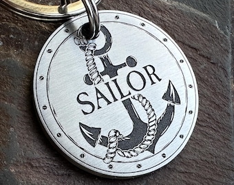 Puede incluir: Un llavero de metal plateado con temática náutica. La etiqueta presenta un diseño de ancla con la palabra "SAILOR" en negrita. El ancla está detallada con cuerda, y la etiqueta tiene un borde circular con pequeños remaches.