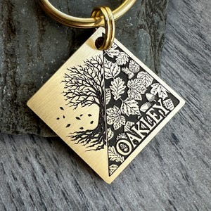 May include: A gold-colored, diamond-shaped keychain tag with a split design. One side features a detailed tree illustration, the other side has leaves and the name "OAKLEY". A gold ring is attached at the top.