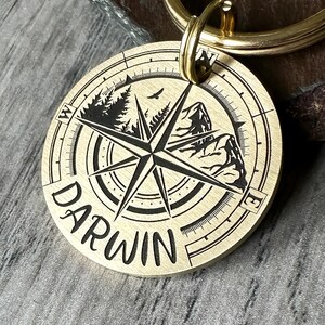 Könnte beinhalten: Ein runder, messingfarbener Schlüsselanhänger mit Kompassdesign. Der Anhänger zeigt eine Windrose, Berge, Bäume und einen Vogel. Das Wort "DARWIN" ist auf dem Anhänger eingraviert. Ein goldfarbener Ring ist am Anhänger befestigt.