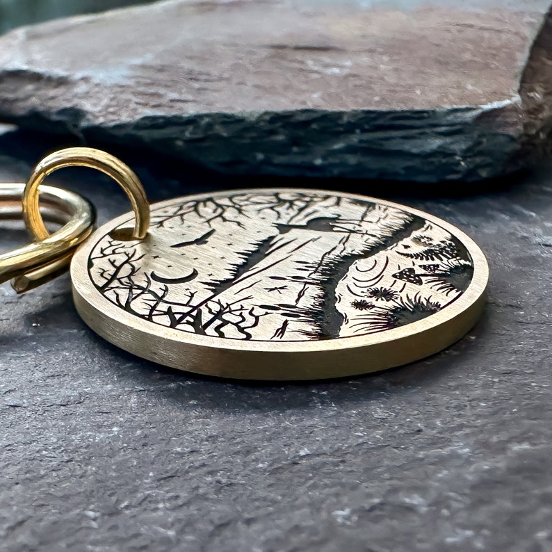 May include: A brass keychain with a detailed engraved design of a forest scene at night. The scene includes trees, a mountain, a crescent moon, and a flying bird. The keychain has a gold-colored ring.
