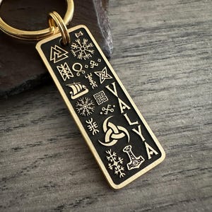 Op de afbeelding: Een rechthoekige goudkleurige metalen sleutelhanger met een zwart verdiept middenstuk, gegraveerd met diverse Noorse symbolen zoals een Valknut, Vegvisir en een Vikingschip, naast de verticale tekst VALYA en een afbeelding van Mjolnir.