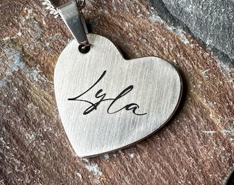 May include: A heart-shaped pendant necklace with a brushed silver-tone finish. The surface features the name Lyla engraved in a black cursive script. The pendant is attached to a matching silver-tone metal chain link necklace.