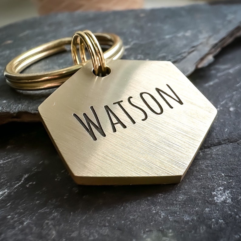 May include: A gold hexagonal dog tag with the name "WATSON" engraved on it. The tag is attached to a gold key ring.