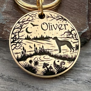 May include: A gold-toned metal dog tag with a black silhouette of a dog standing in a forest scene. The tag is engraved with the name "Oliver" and features a crescent moon, stars, and other woodland details.