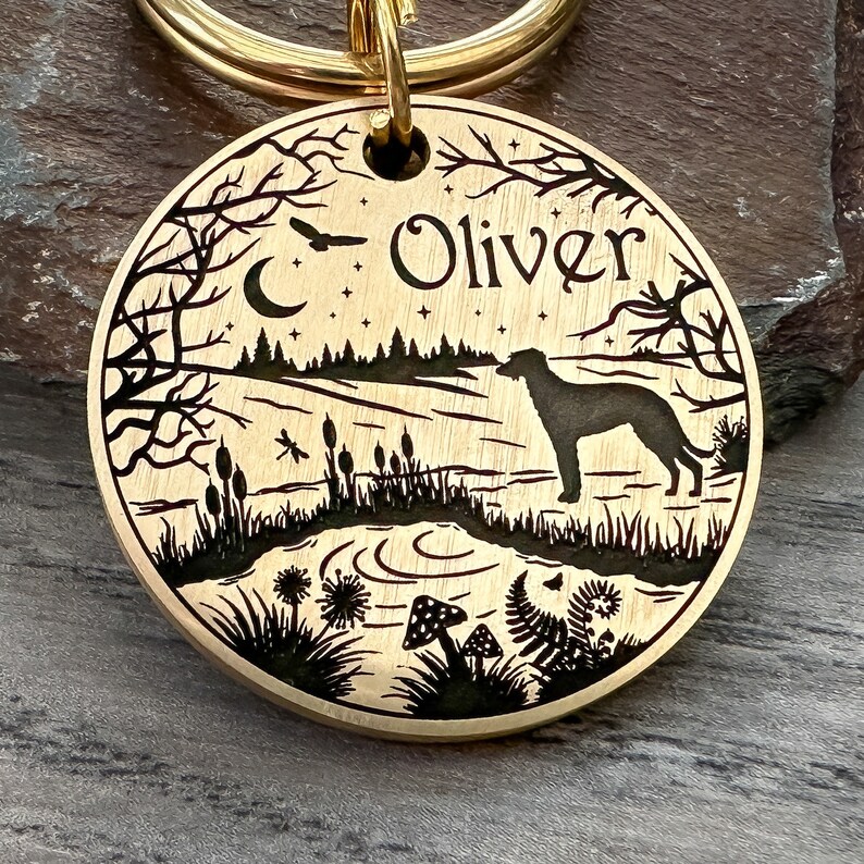 May include: A gold-toned metal dog tag with a black silhouette of a dog standing in a forest scene. The tag is engraved with the name "Oliver" and features a crescent moon, stars, and other woodland details.