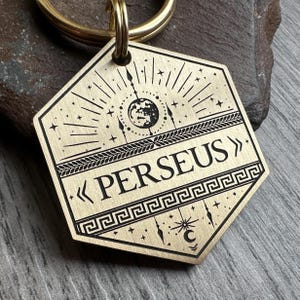 May include: A brass key chain with a hexagonal tag featuring a stylized design of a crescent moon, stars, and a sun with a face. The tag is engraved with the text "< PERSEUS >" and a Greek key pattern.
