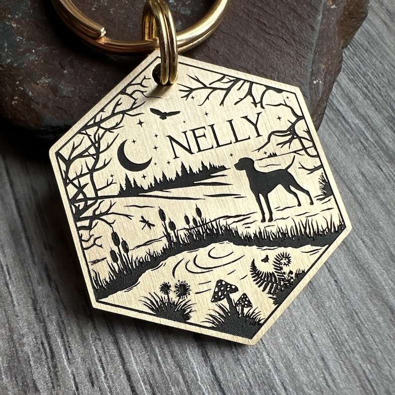 May include: Brass dog tag with a black silhouette of a dog, a crescent moon, and trees. The tag is engraved with the name "NELLY".