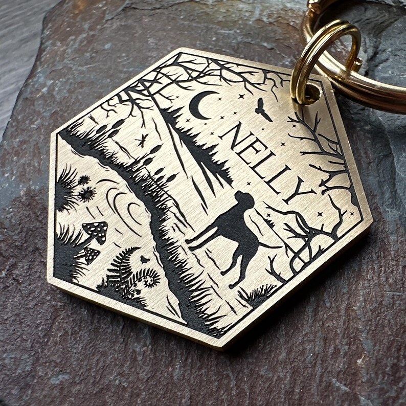 May include: Brass dog tag with a black silhouette of a dog running through a forest scene with a crescent moon and stars. The tag is engraved with the name "NELLY".