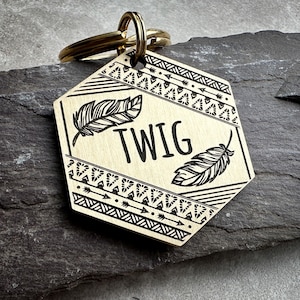 Op de afbeelding: Een zeshoekige, messingkleurige metalen huisdierpenning met een goudkleurige ring. De penning is in het midden gegraveerd met de naam TWIG, geflankeerd door twee gedetailleerde veerillustraties en omrand door geometrische patronen van pijlen en driehoeken.