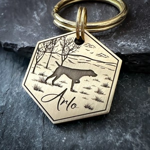 May include: Brass dog tag with a hexagonal shape, featuring an engraved silhouette of a dog in a field with trees and the name "Arlo" inscribed below.