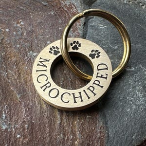 Może przedstawiać: Metalowy, złoty brelok dla psa z wygrawerowanym napisem "MICROCHIPPED" wokół krawędzi. Na breloku wygrawerowane są również trzy odciski łap.