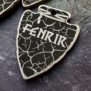 Könnte beinhalten: Schwarzer Holzpfeilspitz mit einem rissigen Oberflächenmuster und dem Wort "Fenrir" in weißen Buchstaben.