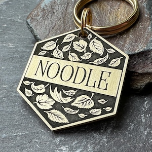 Puede incluir: Chapa de perro de lat&oacute;n con forma hexagonal y un dise&ntilde;o de hojas en blanco y negro. La chapa tiene la palabra "NOODLE" grabada.