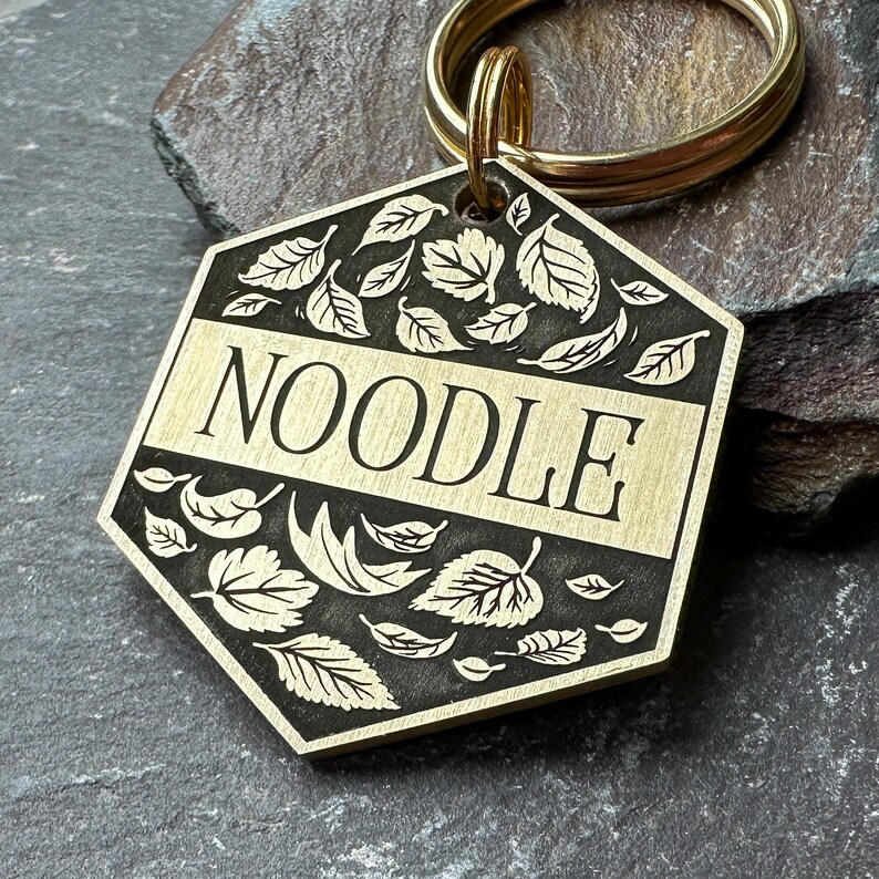 Puede incluir: Chapa de perro de lat&oacute;n con forma hexagonal y un dise&ntilde;o de hojas en blanco y negro. La chapa tiene la palabra "NOODLE" grabada.