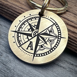 Op de afbeelding: Een messing kompas sleutelhanger met een zwart kompas ontwerp en de letter "A" gegraveerd in het midden. Het kompas is omgeven door een decoratieve rand.