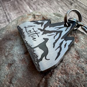 Op de afbeelding: Een zilveren metalen hondenpenning met een berg silhouet en een hond die er vooruit rent. De penning is gegraveerd met de naam "Lottie".