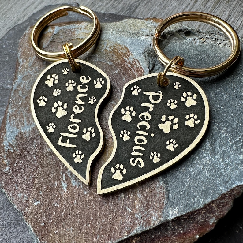 May include: Two gold-tone keyrings with split heart-shaped charms. The charms are black with gold paw prints and the names "Florence" and "Precious" engraved on them.