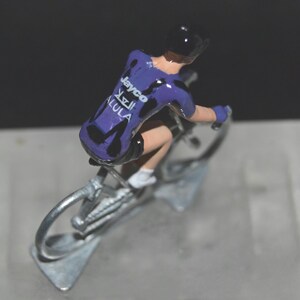 May include: A miniature figurine of a cyclist on a bicycle. The cyclist wears a purple jersey with black accents and the text "Jayco AlUla". The bicycle and cyclist are silver in color.