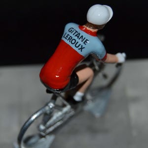 May include: A miniature figurine of a cyclist on a bicycle. The cyclist wears a red and light blue jersey with the text "GITANE LEROUX" on the back. The cyclist is wearing a white helmet and gloves. The bicycle is silver.