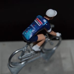 Puede incluir: Una figura de ciclismo azul y blanco con un casco blanco. La figura está montando una bicicleta plateada con un maillot azul y blanco que dice "Roudal" y "Backstep".