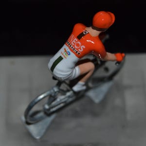Puede incluir: Figura en miniatura de un ciclista con maillot y casco naranja brillante, montado en una bicicleta plateada. El maillot lleva el logotipo de INEOS Grenadiers. El ciclista está en posición de carrera, con las ruedas y el cuadro de la bicicleta visibles.