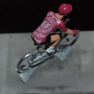 Peut inclure: Figurine miniature d'un cycliste en position de course. Le cycliste porte un maillot rose avec des détails blancs et un casque assorti. Le vélo est de couleur argentée et la figurine est montée sur une base grise.