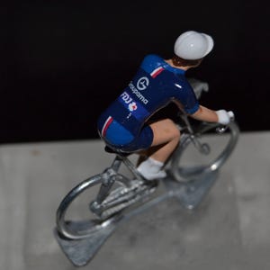 May include: A blue and white cycling figurine of a cyclist riding a silver bicycle. The cyclist is wearing a white helmet and a blue jersey with the words "Groupama FDJ" on it.