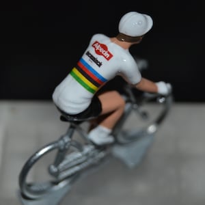 Puede incluir: Una figura blanca de un ciclista que lleva un maillot blanco con las palabras "Alpecin-Deceuninck" y las rayas arcoíris del campeón del mundo. El ciclista va en una bicicleta plateada.
