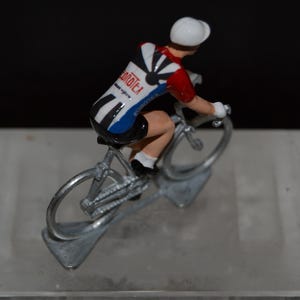 May include: A small figurine of a cyclist on a bicycle. The cyclist wears a white helmet, red and black jersey with the word "onotex" and blue shorts. The bicycle is silver-colored and appears to be made of metal.