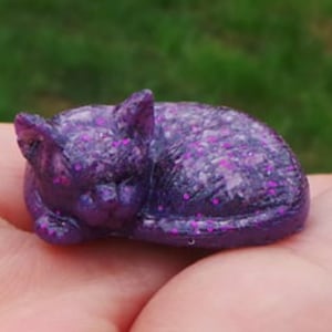 May include: A small purple cat figurine with pink glitter accents. The cat is curled up in a sleeping position.