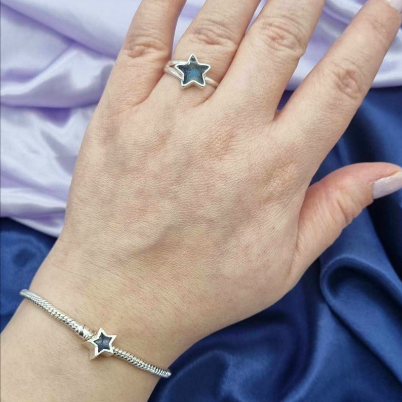 K&ouml;nnte beinhalten: Ein silbernes Armband mit einem schwarzen Stern-Anh&auml;nger und ein silberner Ring mit einem schwarzen Stern. Der Schmuck befindet sich am Handgelenk und Finger einer Person.