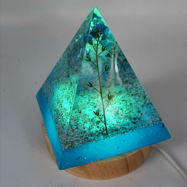 May include: A blue glass pyramid lamp with a small flower pressed inside. The lamp is lit from the inside, creating a soft glow. The base of the lamp is made of wood.