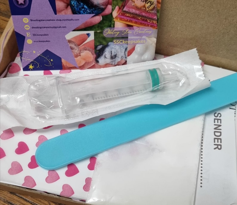 May include: A close-up of a clear plastic syringe with a green tip, a blue nail file, and a white paper with the word "SENDER" printed on it. The items are on a white background with pink hearts.