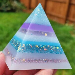 May include: A multi-layered pyramid made of clear resin with blue, purple, and white layers. The resin is filled with iridescent glitter.