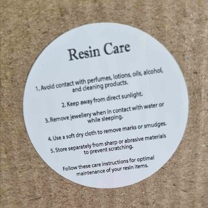 K&ouml;nnte beinhalten: Wei&szlig;er runder Aufkleber mit dem Text "Resin Care" und einer Liste von Pflegehinweisen f&uuml;r Harzgegenst&auml;nde. Die Hinweise beinhalten die Vermeidung von Kontakt mit Parf&uuml;ms, Lotionen, &Ouml;len, Alkohol und Reinigungsmitteln; die Aufbewahrung der Gegenst&auml;nde vor direkter Sonneneinstrahlung; das Entfernen von Schmuck bei Kontakt mit Wasser oder w&auml;hrend des Schlafens; die Verwendung eines weichen, trockenen Tuchs zum Entfernen von Flecken oder Schlieren; und die getrennte Aufbewahrung der Gegenst&auml;nde von scharfen oder scheuernden Materialien, um Kratzer zu vermeiden.