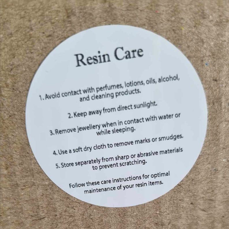 K&ouml;nnte beinhalten: Wei&szlig;er runder Aufkleber mit dem Text "Resin Care" und einer Liste von Pflegehinweisen f&uuml;r Harzgegenst&auml;nde. Die Hinweise beinhalten die Vermeidung von Kontakt mit Parf&uuml;ms, Lotionen, &Ouml;len, Alkohol und Reinigungsmitteln; die Aufbewahrung der Gegenst&auml;nde vor direkter Sonneneinstrahlung; das Entfernen von Schmuck bei Kontakt mit Wasser oder w&auml;hrend des Schlafens; die Verwendung eines weichen, trockenen Tuchs zum Entfernen von Flecken oder Schlieren; und die getrennte Aufbewahrung der Gegenst&auml;nde von scharfen oder scheuernden Materialien, um Kratzer zu vermeiden.