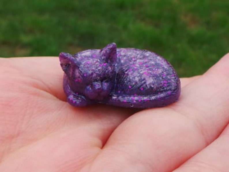 May include: A small purple cat figurine with pink glitter accents. The cat is curled up in a sleeping position.