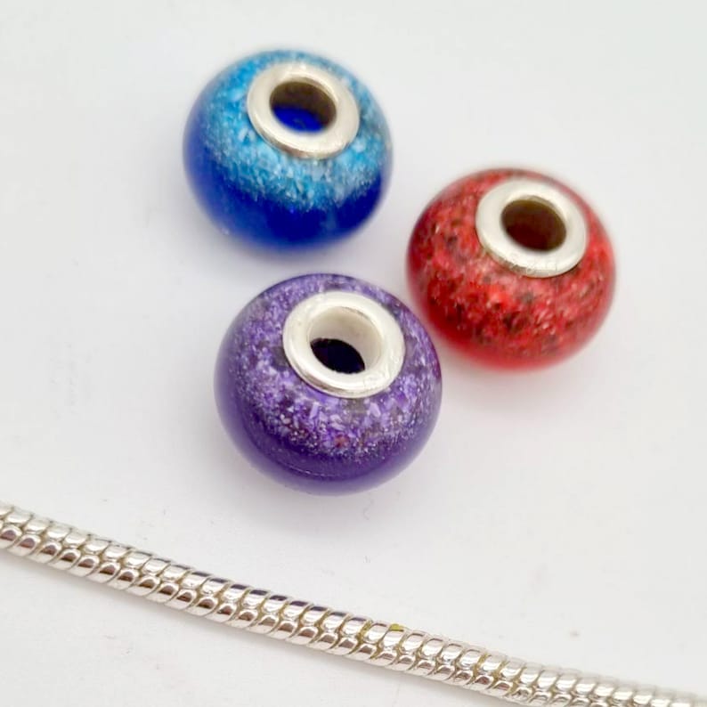 May include: Three glass beads with silver accents. The beads are blue, red, and purple with a speckled pattern. The beads are for jewelry making.