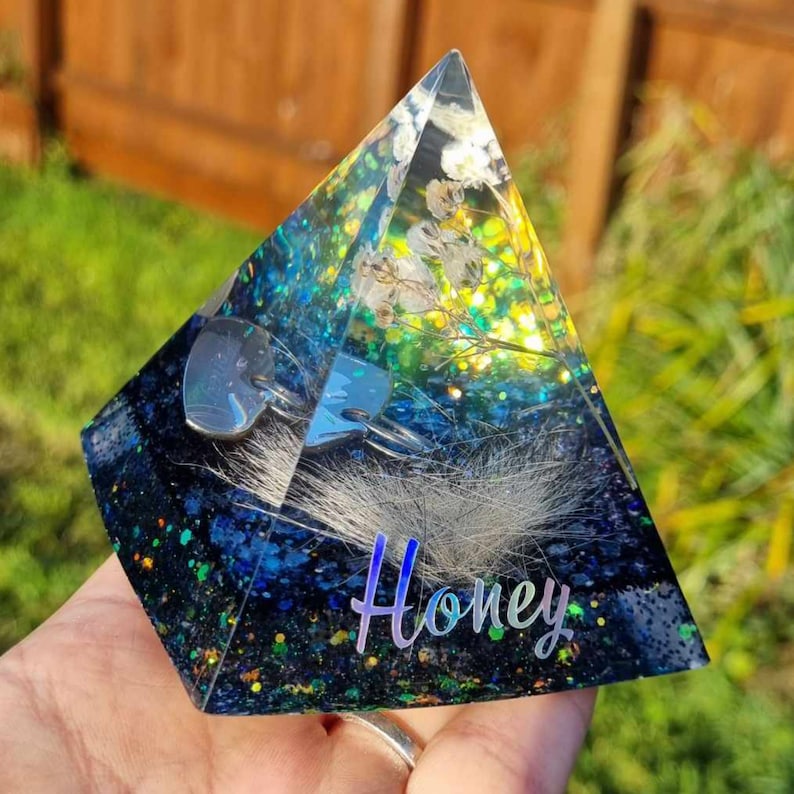May include: A clear glass pyramid filled with glitter, dried flowers, and white fur. The pyramid has a blue and green iridescent base with the word "Honey" written in white.
