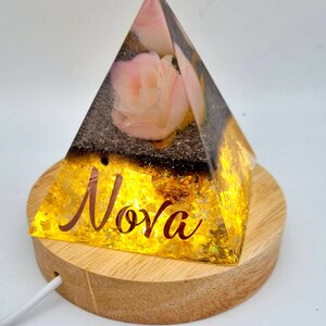 May include: A clear glass pyramid with a pink rose inside, sitting on a wooden base. The pyramid is illuminated from within, casting a warm glow. The base has the word "Nova" inscribed on it.