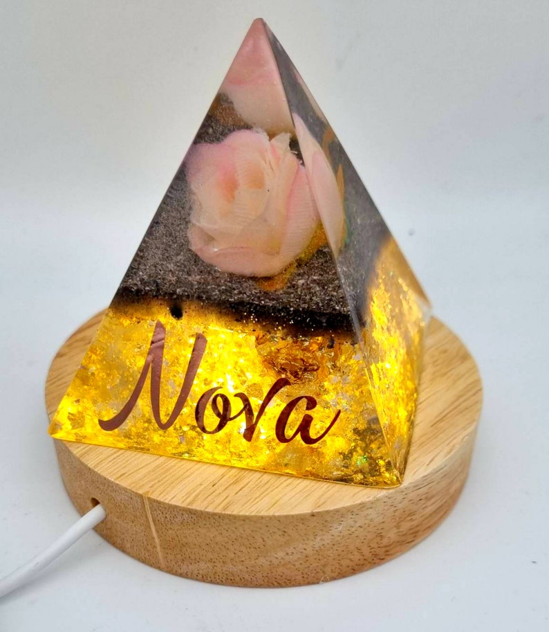 May include: A clear glass pyramid with a pink rose inside, sitting on a wooden base. The pyramid is illuminated from within, casting a warm glow. The base has the word "Nova" inscribed on it.