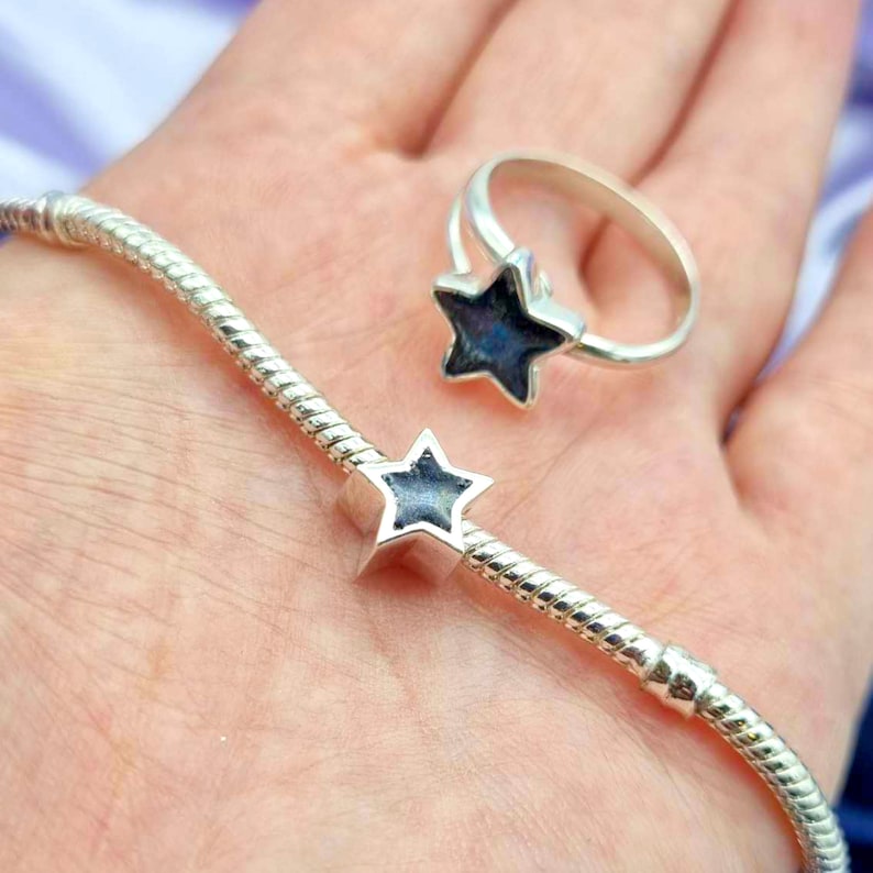 K&ouml;nnte beinhalten: Ein silbernes Armband mit einem sternf&ouml;rmigen Anh&auml;nger. Der Anh&auml;nger hat eine dunkelblaue Mitte und einen silbernen Umriss. Das Armband befindet sich am Handgelenk einer Person.