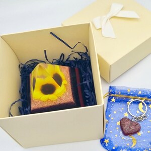 May include: A clear glass pyramid with a sunflower design inside. The pyramid is in a white box with tissue paper and a blue and gold patterned bag with a heart-shaped charm.