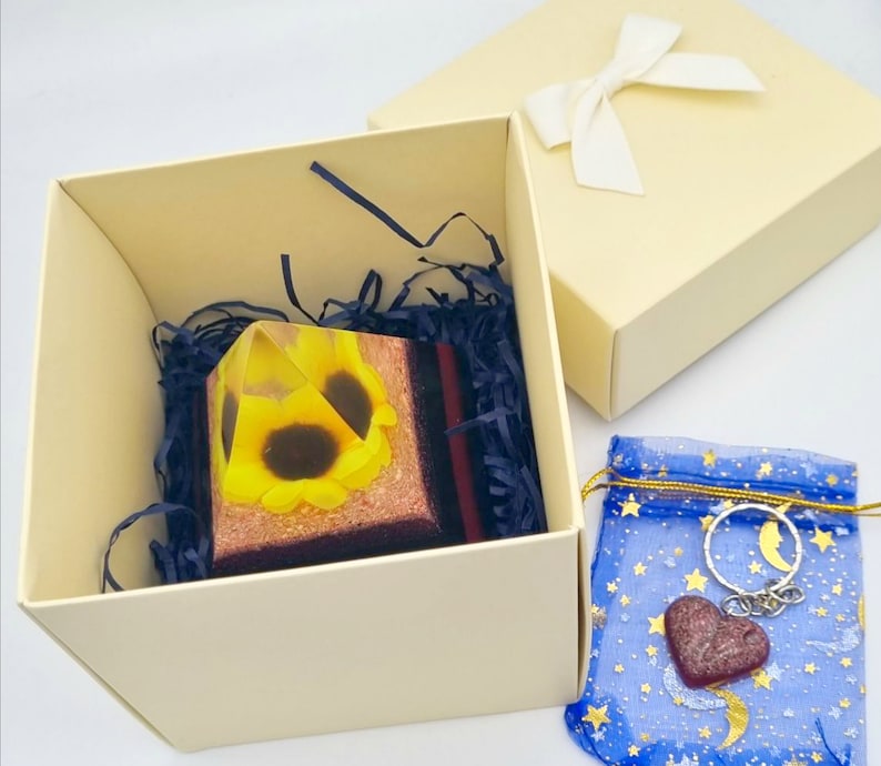 May include: A clear glass pyramid with a sunflower design inside. The pyramid is in a white box with tissue paper and a blue and gold patterned bag with a heart-shaped charm.