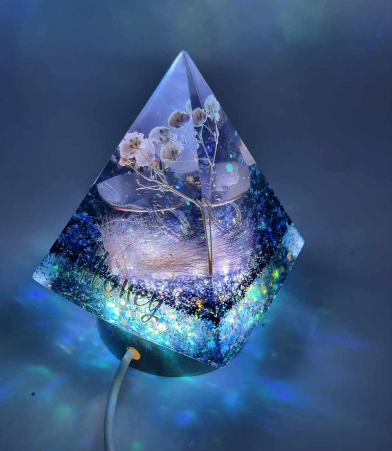 May include: A blue resin pyramid with a white flower and glitter inside. The pyramid is lit from the bottom with a white cord.