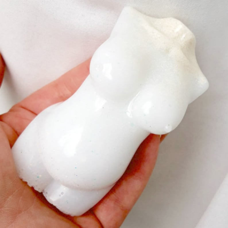 May include: White ceramic sculpture of a female torso with a smooth, glossy finish. The sculpture has a subtle sparkle.
