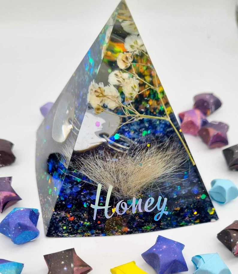 May include: A clear glass pyramid filled with dried flowers, glitter, and a small piece of white fur. The word "Honey" is written on the base of the pyramid.