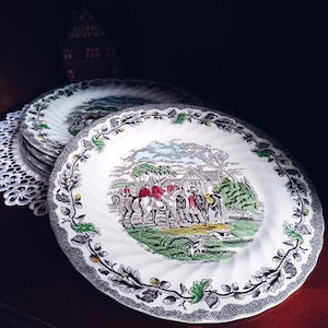Pode incluir: Uma pilha de pratos de cerâmica branca com uma impressão central de uma cena de caça à raposa com cavaleiros em cavalos e cães. A borda é decorada com um padrão de folhas de carvalho e bolotas em preto e branco com detalhes verdes.