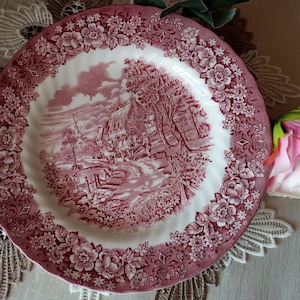 Può includere: Un piatto rotondo in stile antico con un design floreale e paesaggistico rosso e bianco. Il piatto presenta un'illustrazione dettagliata di una scena rurale con una casa, alberi e una strada tortuosa. Il bordo è decorato con un motivo floreale.
